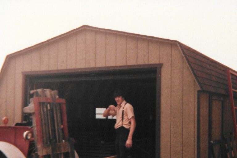 A man standing at the entrance of a barn-style shed, giving a thumbs-up gesture. In the foreground, a forklift is carrying long wooden planks. The scene includes various pieces of farm equipment and spare tires stacked beside the shed. The shed has a peaked roof and sliding doors, with a red metal gate leaning against the side.