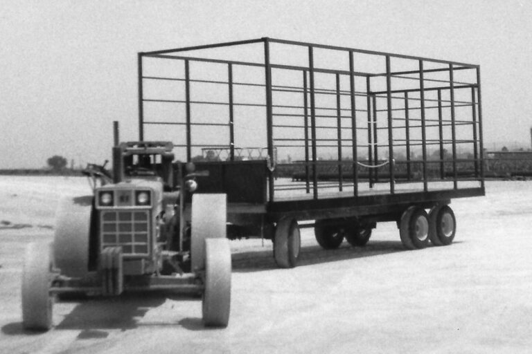 Black-and-white image of a tractor pulling a large, open metal frame structure on a flatbed trailer. The frame appears to be a large livestock or equipment cage. The scene is set on a farm with open land in the background and a silo and other farm buildings visible in the distance