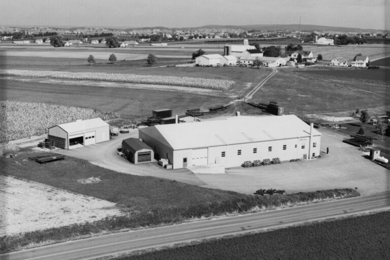 Aerial black-and-white image of a large industrial facility surrounded by farmland. The main building has a flat roof, and several smaller outbuildings are nearby. Large stacks of materials and equipment are arranged around the site. In the distance, fields, farmhouses, and silos are visible, stretching across the rural landscape.