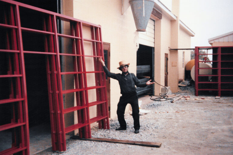 A man wearing a straw hat and dark clothing standing outside a building, gesturing towards large red metal gates leaned against the wall. The area around him has gravel, some equipment, and an industrial structure in the background