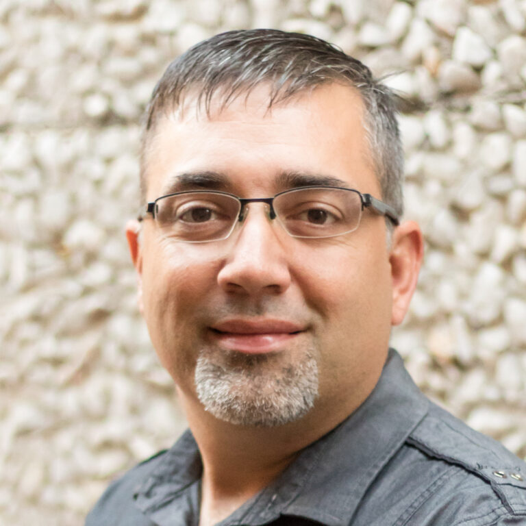 A man with short dark hair, glasses, and a trimmed goatee, wearing a dark gray collared shirt, standing in front of a textured stone wall