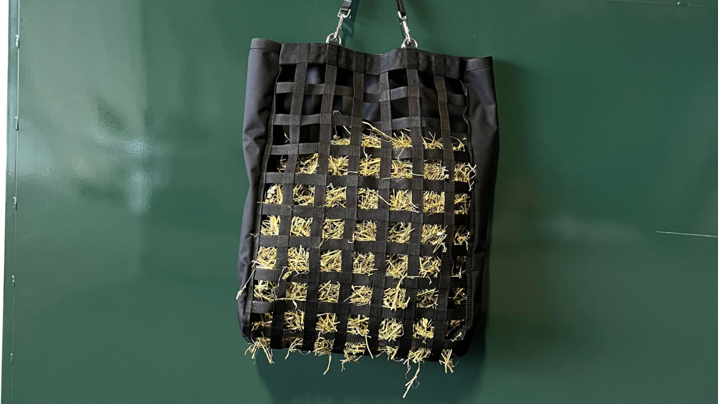 A close-up of a black hay bag feeder with mesh webbing filled with hay. The bag is suspended from two metal clips, attached at the top. The mesh design allows animals to pull small amounts of hay through the squares, reducing waste. The background is a smooth, dark green wall, creating a clean and simple backdrop that emphasizes the feeder.