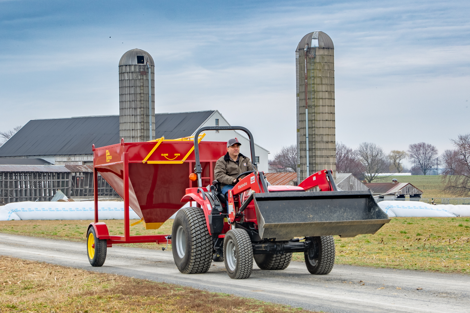 A farmer drives a red tractor down a dirt road, transporting a large portable grain bin attached to the back of the tractor. The bright red bin contrasts with the overcast sky and muted colors of the surrounding farm buildings, silos, and fields. The grain bin, which is used for feeding livestock, is on yellow wheels and labeled with an easy-to-read "Close" and "Open" mechanism. The farmer, dressed in a brown jacket and gray cap, appears focused as he navigates the equipment along the rural farm path, framed by silos and agricultural buildings in the background.